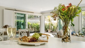 Una mesa con una tabla de quesos, uvas, nueces y una botella de vino. Hay un jarrón con flores y vistas de un jardín a través de grandes ventanales.
