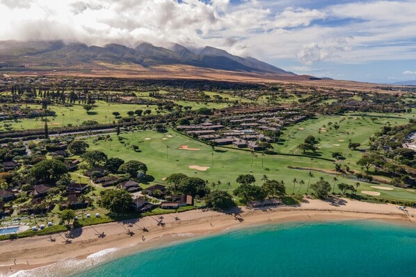 An aerial view of a coastal resort with a golf course, beachfront, and mountainous backdrop.