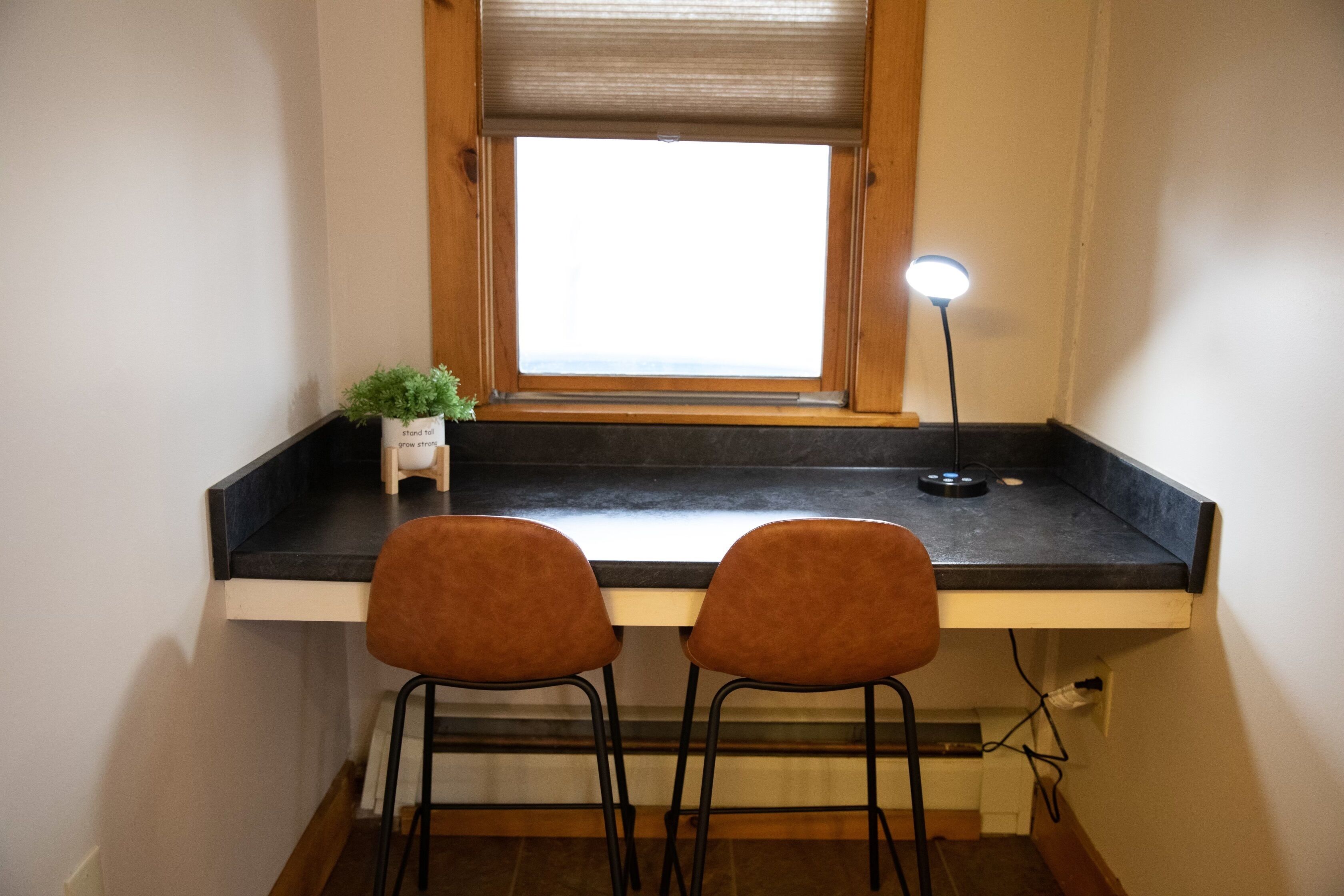 Une petite cuisine avec une fenêtre, deux tabourets, une plante en pot et un lampadaire.