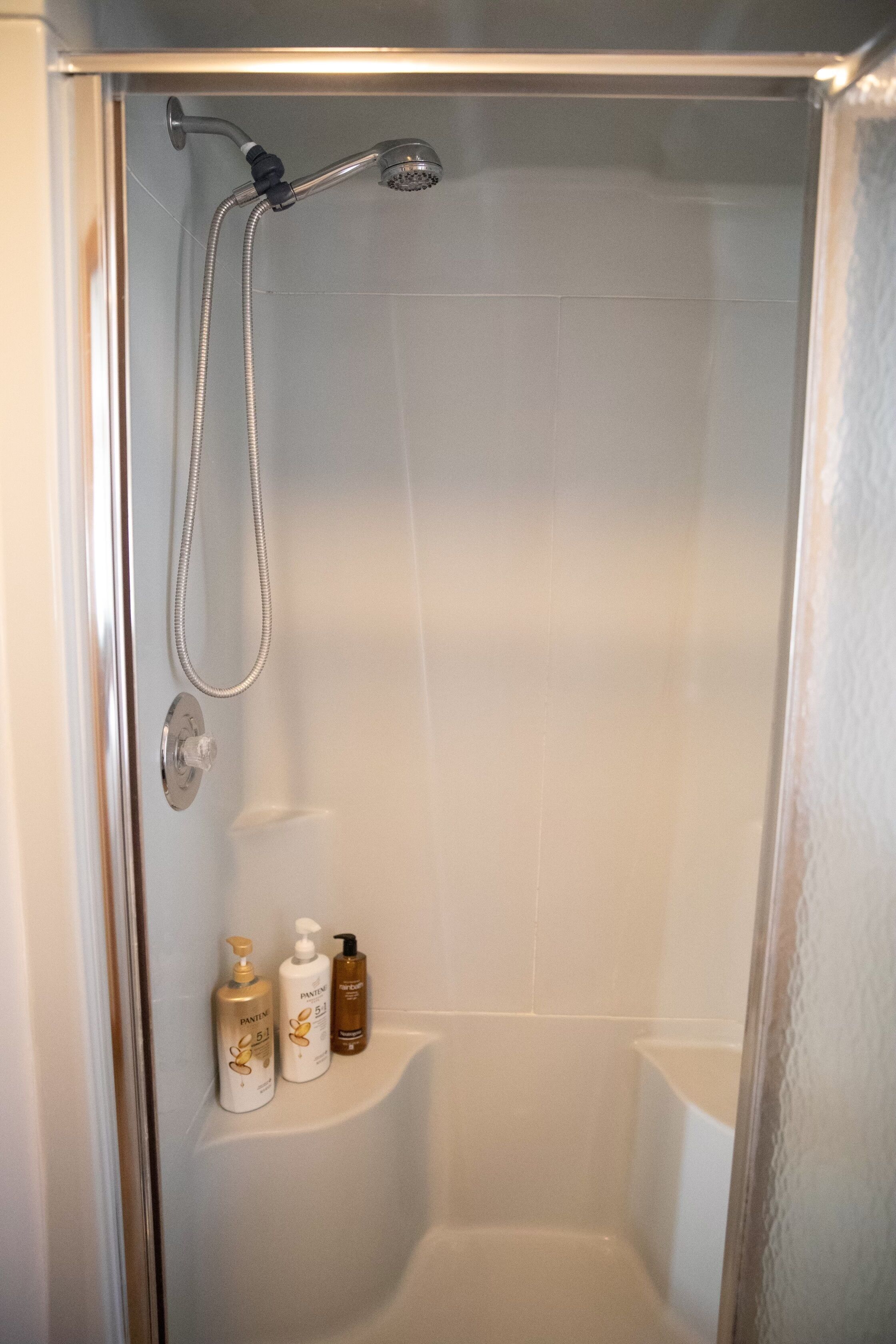 A shower with a handheld showerhead and a handheld hose. There are three bottles of shampoo and conditioner on the wall.