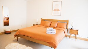 A bedroom with a wooden bed, orange bedding, a white rug, a bedside table with a lamp, and a framed picture on the wall. - Charming 1-bedroom apartment in Jericho  (Jericho)