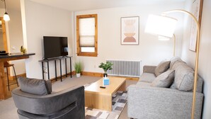 A modern living room with a sofa, a coffee table, a TV, a dining area with a bar, and a window with blinds. - Charming 1-bedroom apartment in Jericho  (Jericho)