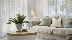Una sala de estar con un sofá beige, una mesa redonda de madera, un helecho en maceta y una lámpara al fondo.