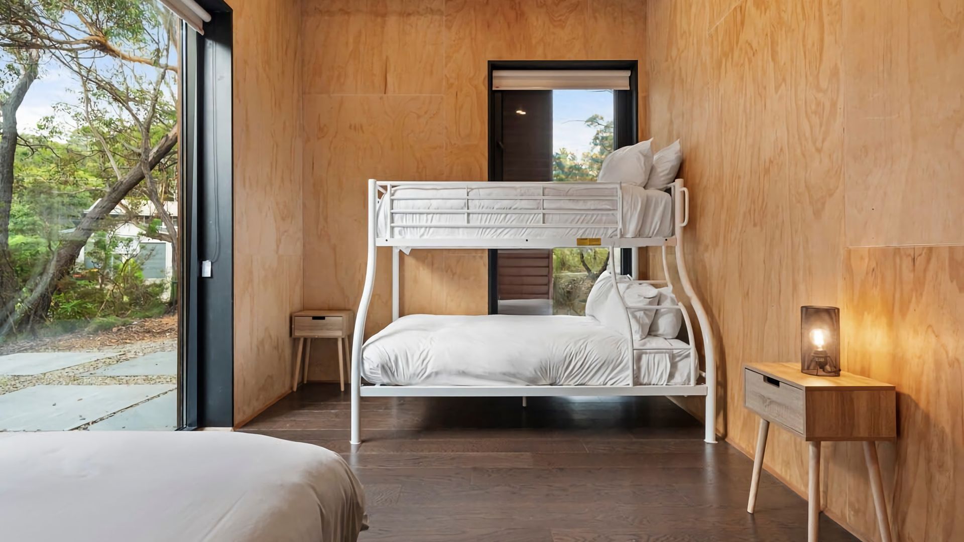 Een slaapkamer met stapelbed, een houten vloer, een nachtkastje met lamp en uitzicht op bomen.
