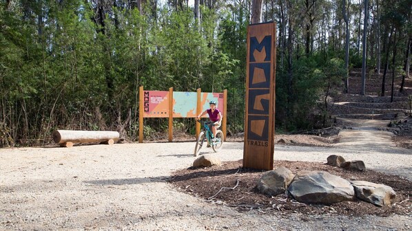 A mountain biking trail entrance with a wooden sign, a cyclist, and a map board in a forested area.