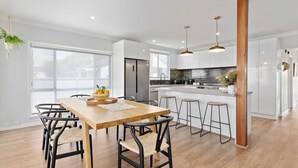 A modern kitchen with a wooden dining table, bar stools, and a large window providing natural light. - Sip and Soak Up Summer in the Hunter (Cessnock)