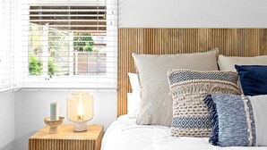 A bedroom with a bed, pillows, a bedside table with a lamp, and a window with blinds. - Sunset Splash Retreat (Budgewoi)