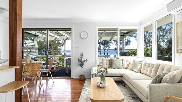 A modern living room with a large window, a sofa, a coffee table, and a dining area with wicker chairs. - Sunset Splash Retreat (Budgewoi)