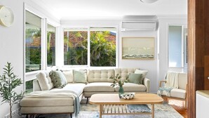 A modern living room with a sectional sofa, a coffee table, and a view of a brick building and greenery through large windows. - Sunset Splash Retreat (Budgewoi)