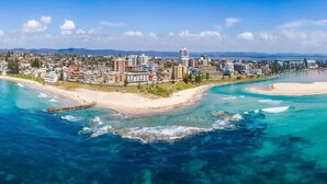 An aerial view of a coastal city with a beach, clear blue waters, and a mix of residential and commercial buildings. - Bobby's Binburra Beach House (Toowoon Bay)