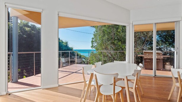 A modern dining area with a large table and chairs, large glass doors leading to a balcony with a view of the sea. - Bobby's Binburra Beach House (Toowoon Bay)