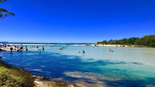 A beach with clear blue water, people swimming and sunbathing, and a distant shoreline with trees.