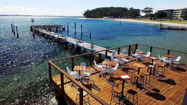 A wooden deck with tables and chairs overlooking a body of water. - Seashells Sweet Retreat Pool, Walk to Shops & Marina (Nelson Bay)