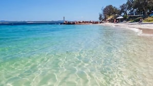 A beach with clear shallow water, a sandy shore, and a few umbrellas. - Seashells Sweet Retreat Pool, Walk to Shops & Marina (Nelson Bay)