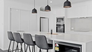 A modern kitchen with white cabinetry, a central island with bar stools, and stainless steel appliances. - Seashells Sweet Retreat Pool, Walk to Shops & Marina (Nelson Bay)