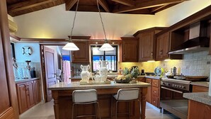 Een keuken met houten kastjes, een centraal kookeiland met barstoelen, een hanglamp en een groot raam.