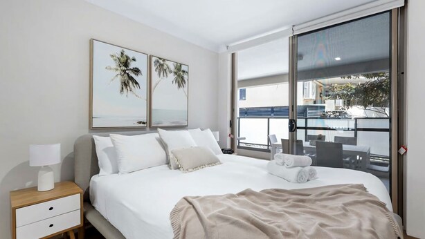 A hotel room with a bed, a nightstand, a lamp, two framed pictures, and a large window with a view of the outside. - Coastal Retreat in the Heart of Kiama (Kiama)