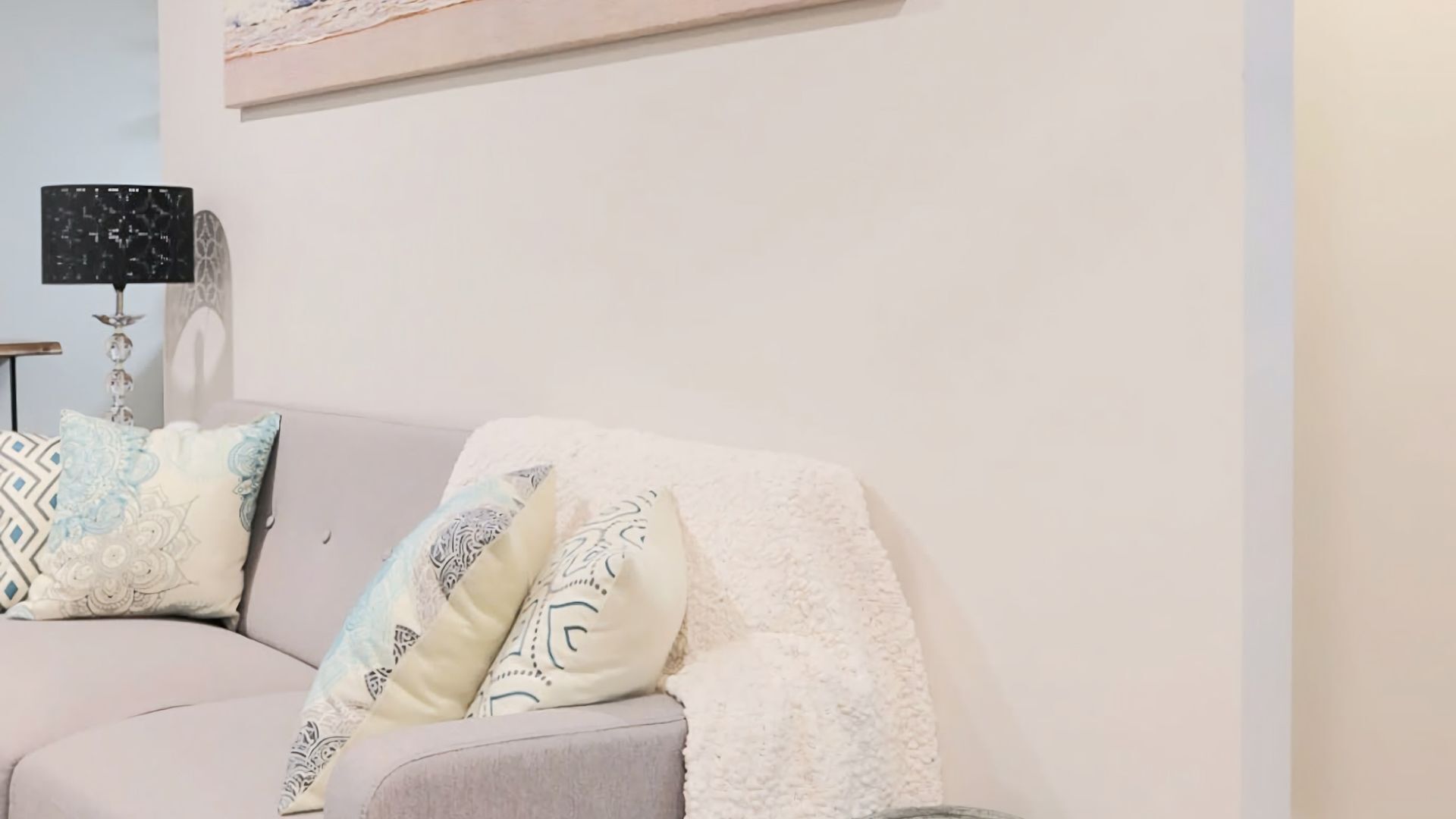 A living room with a sofa, patterned cushions, a decorative lamp, and a wall-mounted shelf.