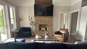 A living room with a stone fireplace, a large flat-screen TV mounted above it, a dark blue sofa, a leather armchair, and a coffee table with decorative items. - Private Heated Pool! @ The House on the Cove (Hiawassee)