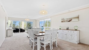 A spacious dining area with a large table and white chairs, a white cabinet with glassware, and large windows offering a view of the outdoors. - Oscar on Main - Heart of Main Beach (Main Beach)