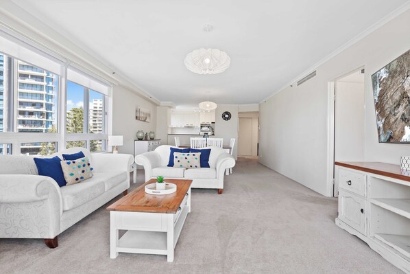 Un salón amplio con mobiliario blanco, una mesa de centro de madera y ventanas grandes con vistas a la ciudad.
