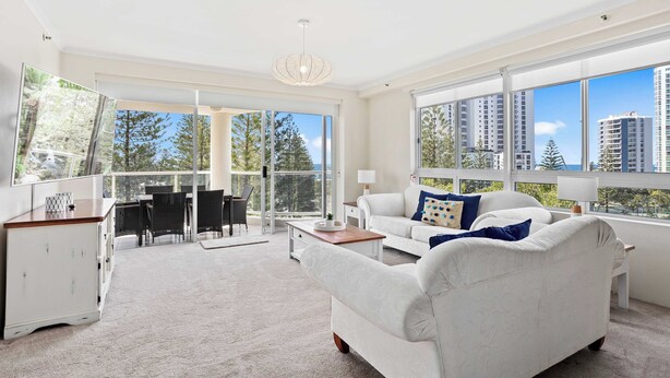 A spacious living room with a large window, a white sofa, a coffee table, and a dining area with chairs and a table. - Oscar on Main - Heart of Main Beach (Main Beach)
