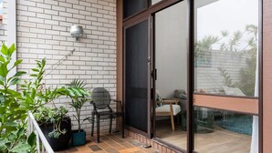 Un balcón con suelo de baldosas, plantas en macetas, una silla y una puerta corrediza de vidrio que da a una zona de piscina.