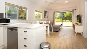 A modern kitchen with white cabinetry, a built-in microwave, and a dishwasher. There is a dining area with a table and chairs, and a large window providing a view of the outdoors. - Broulee Beachfront Bliss - Near the Waves (Broulee)