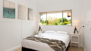 A modern bedroom with a large bed, two bedside tables with lamps, and a window with a view of greenery. - Broulee Beachfront Bliss - Near the Waves (Broulee)