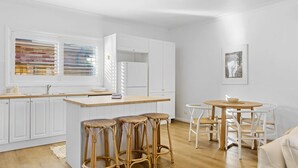 A modern kitchen with white cabinetry, a central island with stools, a dining area with a round table and chairs, and a window with blinds.