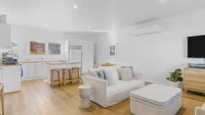 A modern living room with a white sofa, a coffee table, and a TV. There is a kitchen area with white cabinets and a dining table.