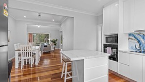 Una cocina moderna con armarios blancos, encimera de mármol y comedor con suelo de madera y sillas blancas.