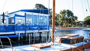 Un barco con cubierta azul y estructura de vidrio, amarrado cerca de una orilla con árboles y un parque infantil al fondo.