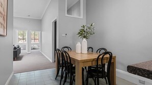 Un coin repas avec une table en bois, des chaises noires et des vases blancs. Une porte donne accès à une autre pièce où se trouvent un canapé et une fenêtre.