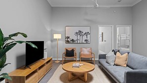 Un salon moderne avec un téléviseur à écran plat, deux fauteuils en bois, une table basse ronde, un canapé gris et une grande plante.