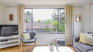 Una sala de estar con televisión, un sofá gris, una mesa de centro blanca y una ventana con cortinas.