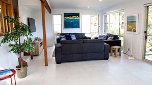 A modern living room with a grey sofa, a wooden coffee table, and a large window with a view of the outdoors. - Blissful Stay Moments From the Beach (Surf Beach)