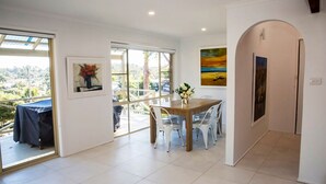 A dining area with a wooden table and white chairs, a painting on the wall, and a view through large windows. - Blissful Stay Moments From the Beach (Surf Beach)