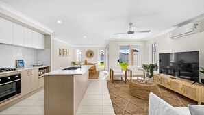 A modern kitchen with white cabinets, a central island, and a dining area with a TV and seating. - Sea La Vie One - Salt Air & Sunsets (Salamander Bay)