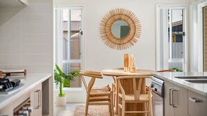 A modern kitchen with a round wooden table, two chairs, a potted plant, and a unique wooden mirror on the wall. - Sea La Vie One - Salt Air & Sunsets (Salamander Bay)