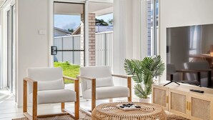 A modern living room with two white armchairs, a round wooden coffee table, a large flat-screen TV, and a potted plant. - Sea La Vie One - Salt Air & Sunsets (Salamander Bay)