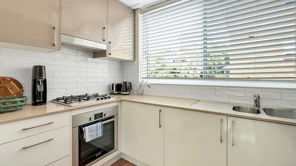 A modern kitchen with white cabinets, a gas stove, a built-in oven, a coffee maker, and a window with blinds.