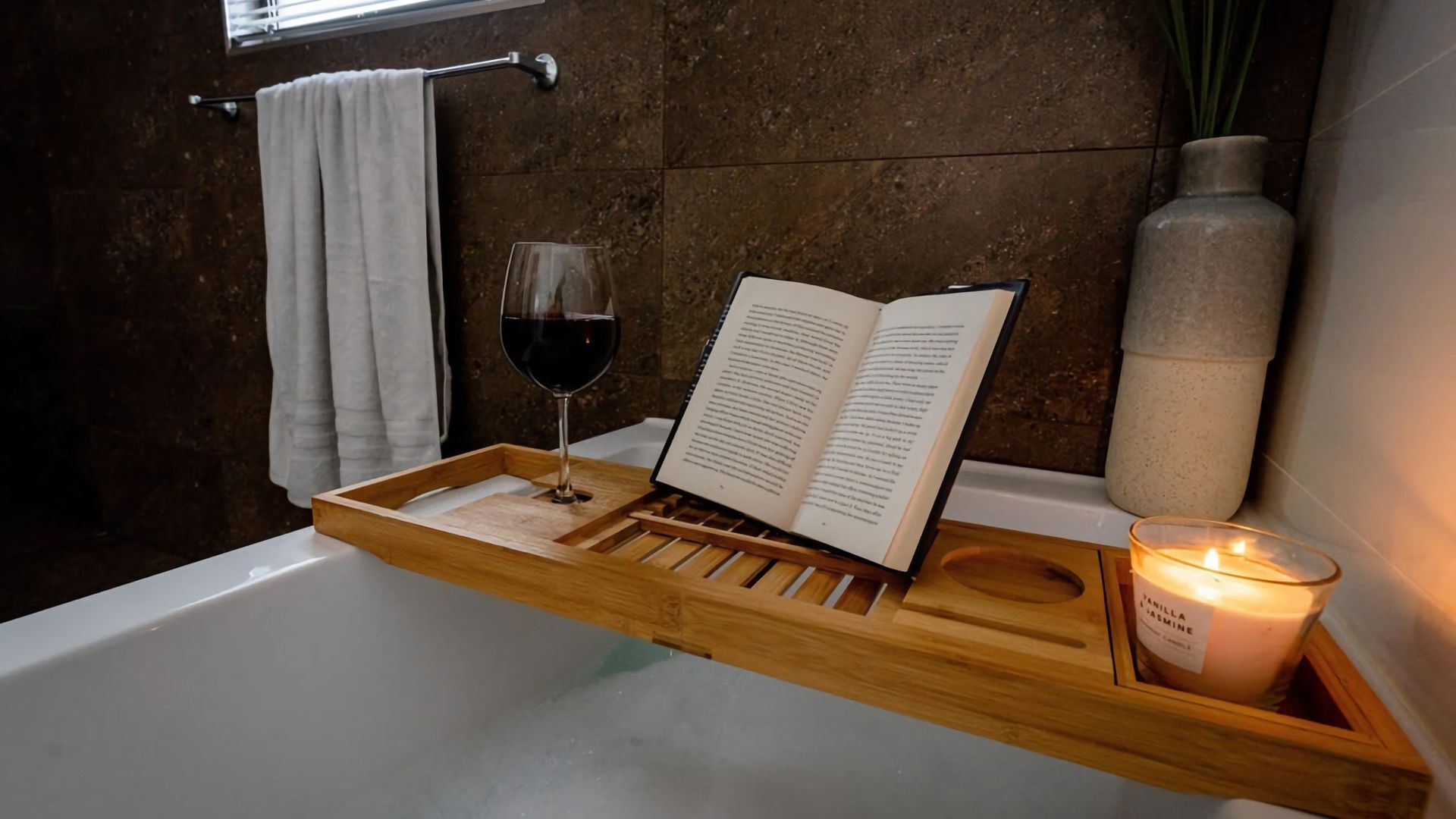 A wooden tray with a book and a glass of red wine on a bathtub.