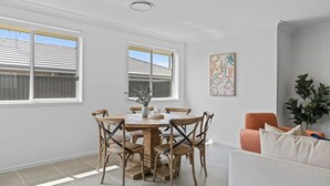 Ein Essbereich mit einem runden Tisch, sechs Stühlen und einer Vase mit Kerzen. Zwei Fenster mit Jalousien, ein Gemälde an der Wand und eine Pflanze in der Ecke sind zu sehen.