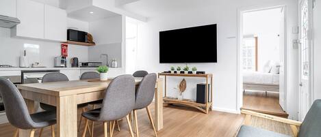 Une cuisine moderne avec des meubles blancs, un four à micro-ondes et une machine à café. On y trouve un coin repas avec une table en bois et des chaises grises. Un téléviseur à écran plat est fixé au mur, au-dessus du coin repas.