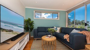 A modern living room with a large TV, a dark blue sofa, a small round table, and a view of the ocean through large windows.