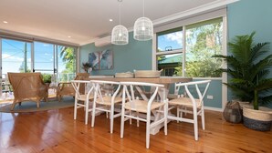 A spacious dining area with a wooden table and white chairs, large windows with a view of greenery, and hanging pendant lights.
