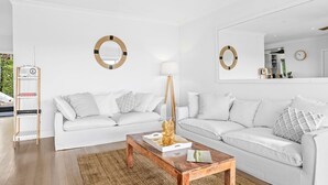 Una sala de estar moderna con un sofá blanco, una mesa de centro de madera y una lámpara de pie. Hay dos espejos montados en la pared y un estante pequeño con libros.