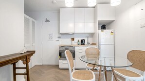 A modern kitchen with white cabinets, a built-in oven, a refrigerator, and a glass dining table with wicker chairs. - The Little Bondi Base (Bondi Junction)