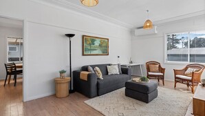 Una sala de estar moderna con un sofá gris, una mesa de centro redonda, sillas de mimbre, un cuadro en la pared y un ventanal con persianas.